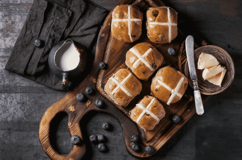 hot cross buns on a wooden board with a cup of butter and a jug of milk